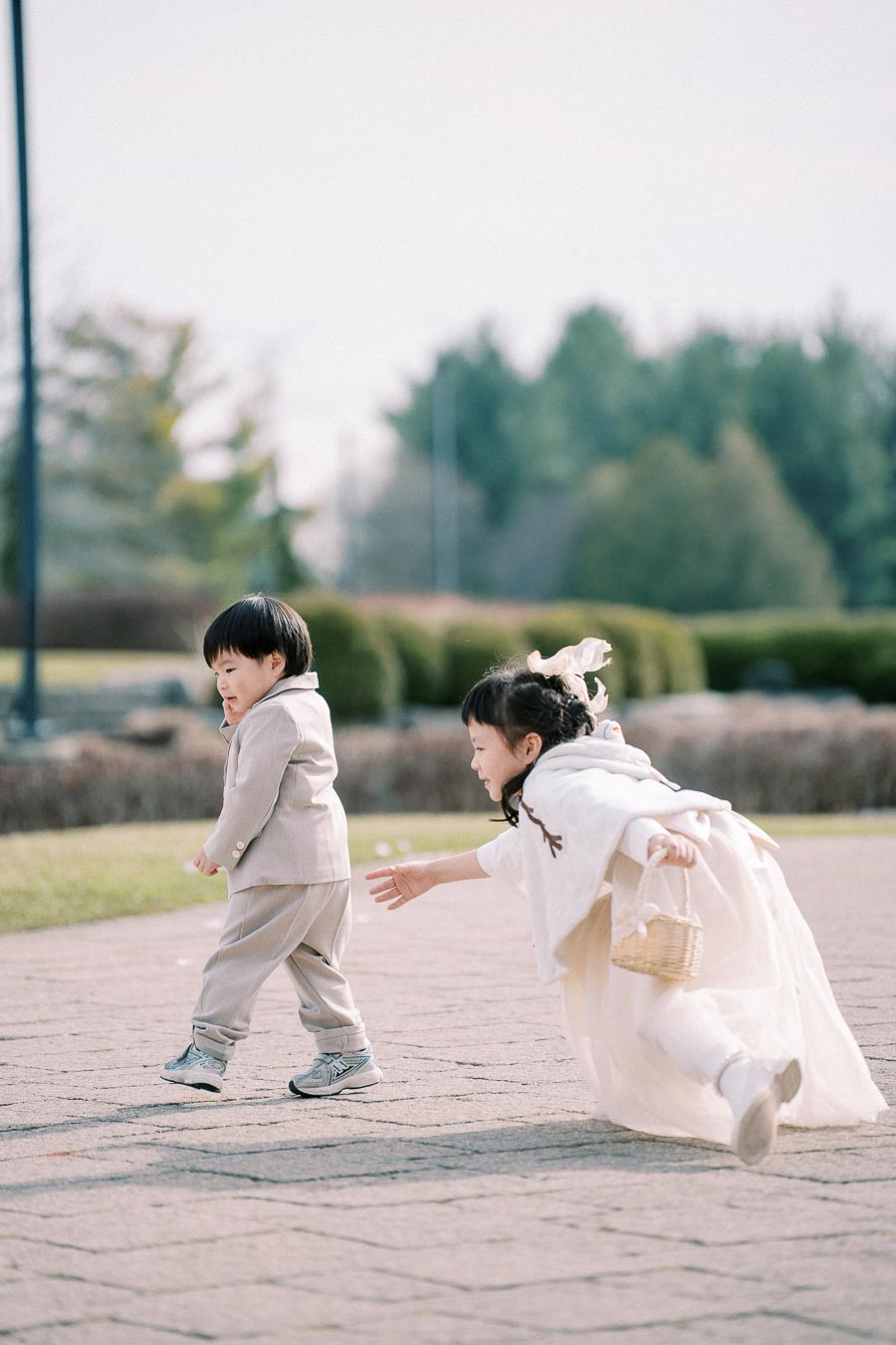 Children playing outdoors on a sunny day, with a girl in a white dress chasing a boy in a light-colored suit on a paved path surrounded by greenery.