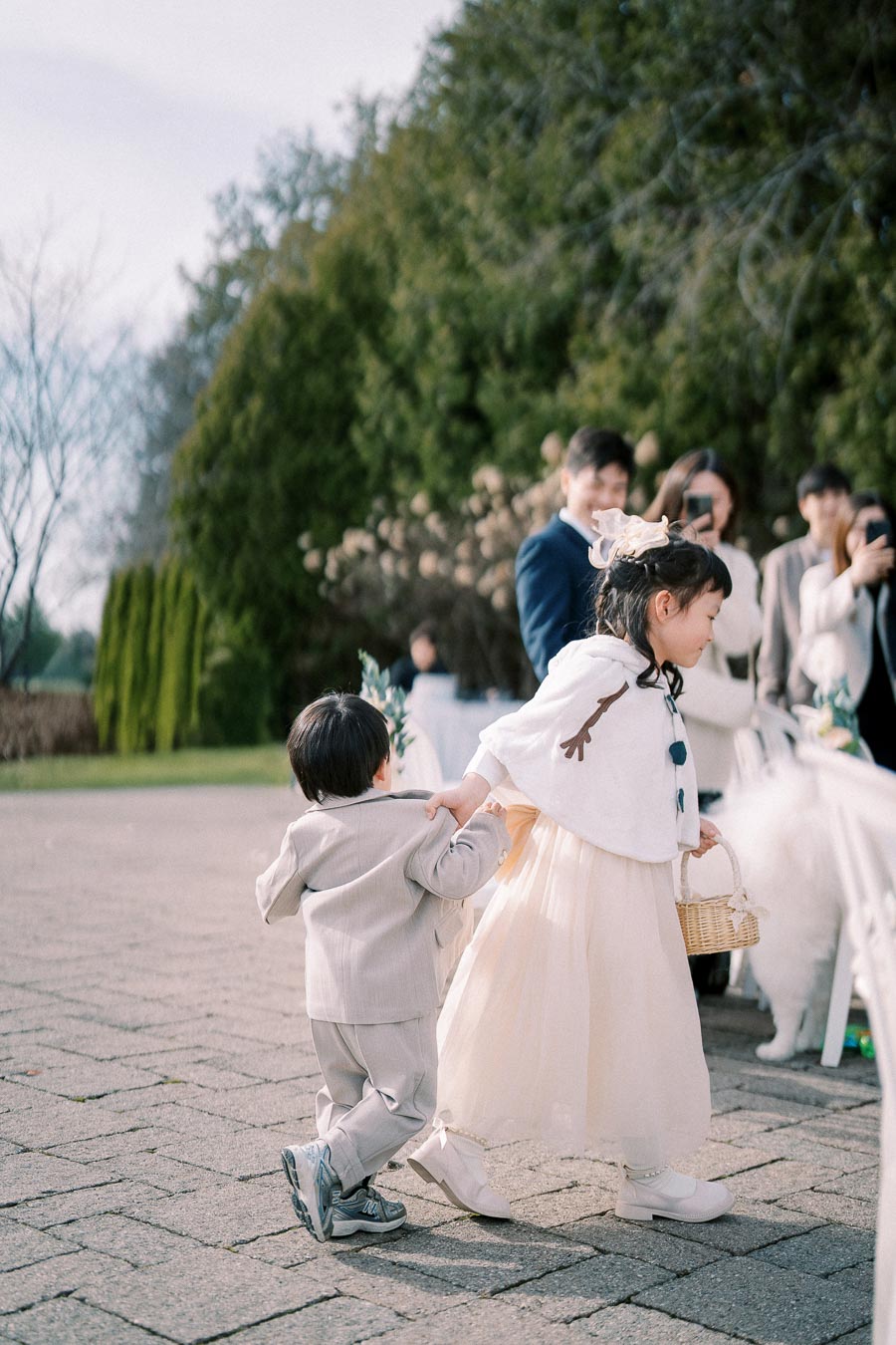 Young flower girl and boy walking together at an outdoor wedding, surrounded by guests in elegant attire, with a lush green background.