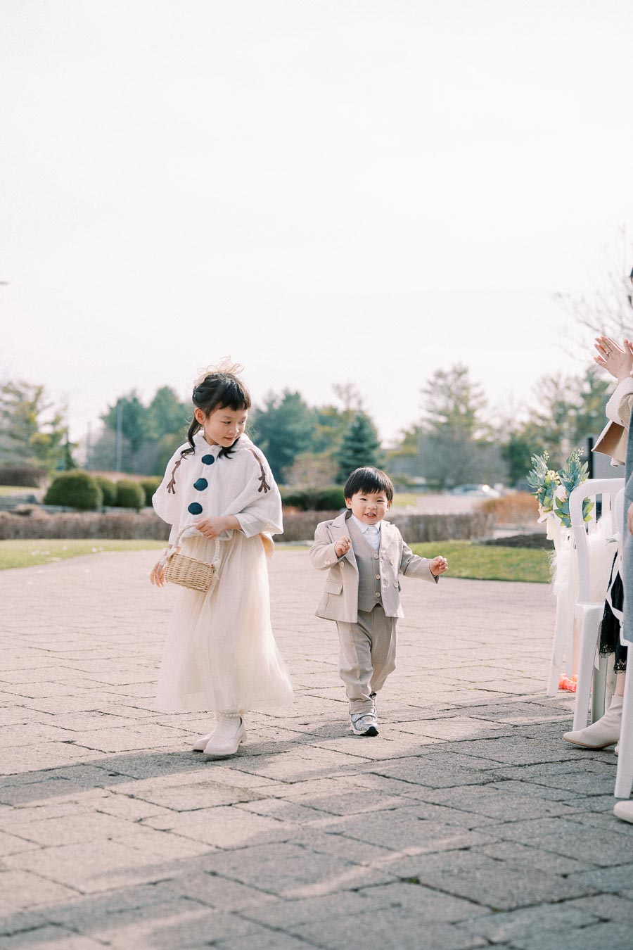 Two young children dressed elegantly, walking outdoors on a paved pathway during a sunny day, with trees in the background. The girl holds a basket and wears a light-colored dress, while the boy is dressed in a light gray suit.