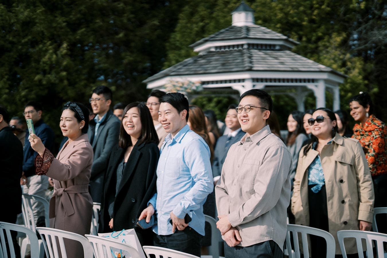 A group of people smiling at an outdoor event with a gazebo in the background, surrounded by green trees.