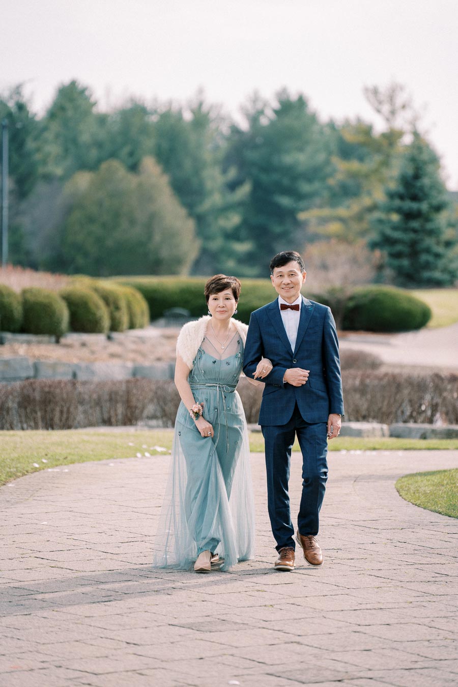 A couple elegantly walking arm in arm outdoors on a paved path, with the woman in a light blue gown and the man in a dark suit with a bow tie, set against a lush green garden backdrop.