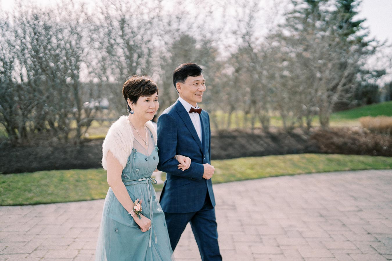 A couple in formal attire walking on a paved path outdoors, with bare trees and grass in the background on a clear day.