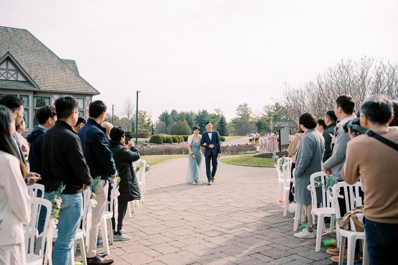 Outdoor wedding ceremony with guests seated on either side of a brick pathway; a couple walks arm-in-arm towards the venue, with greenery and a scenic backdrop under a clear sky.