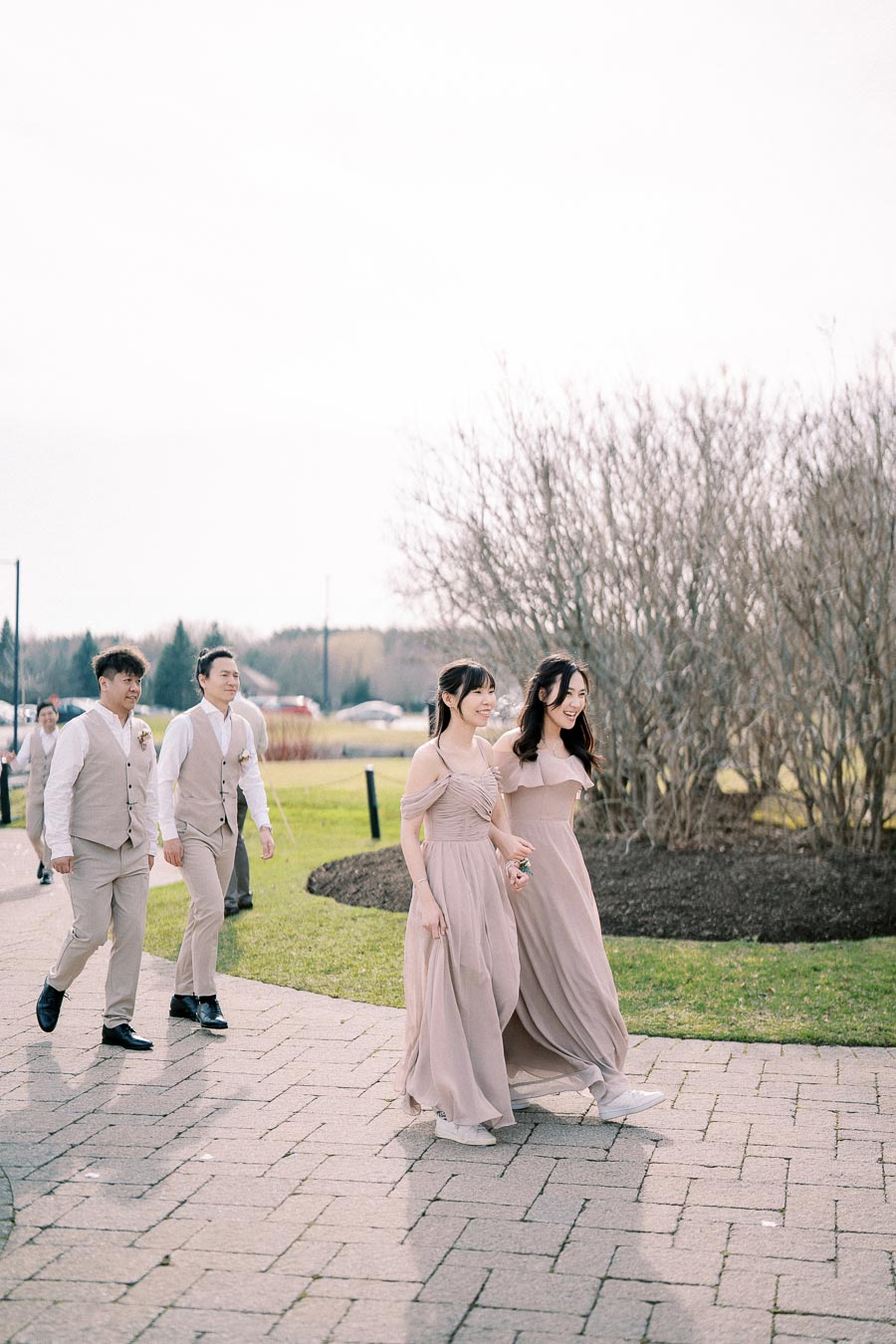 A group of bridesmaids and groomsmen walking outside on a sunny day, dressed in elegant attire, with two women in flowing taupe dresses and two men in light gray suits with vests, conveying a cheerful and formal wedding atmosphere.