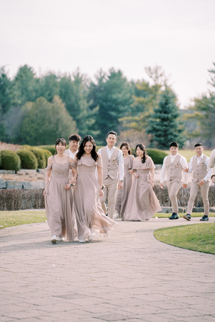 Group of bridesmaids and groomsmen walking down a scenic path during an outdoor wedding ceremony, wearing coordinated light pink dresses and beige suits, surrounded by lush greenery and clear skies.