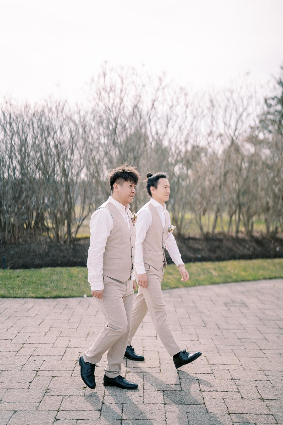 Two groomsmen in coordinating beige vests and pants walk side by side on a paved path, with a background of leafless trees on a clear day.