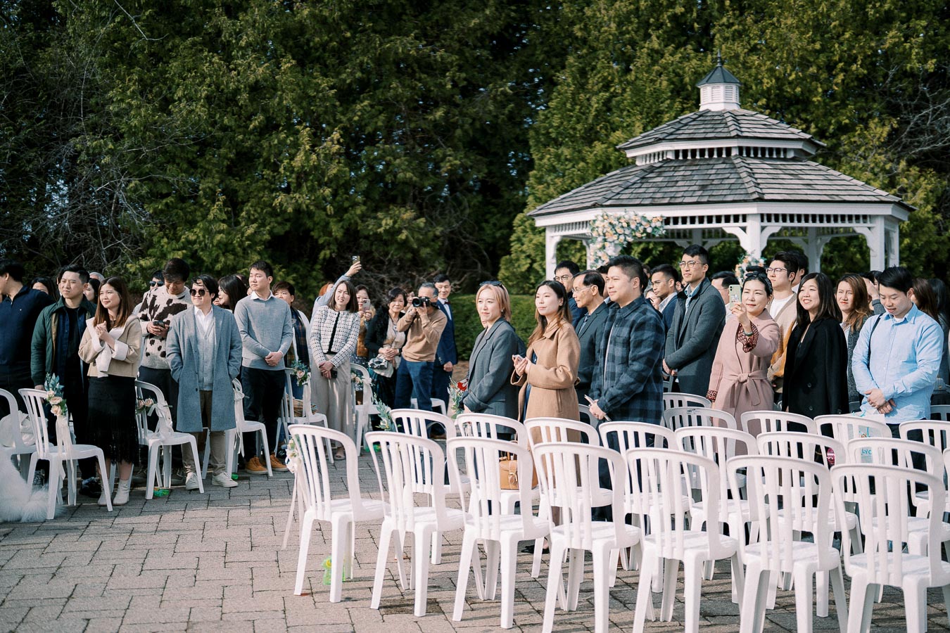 Outdoor event with attendees standing near white plastic chairs, capturing moments on their phones. A gazebo surrounded by greenery is visible in the background.