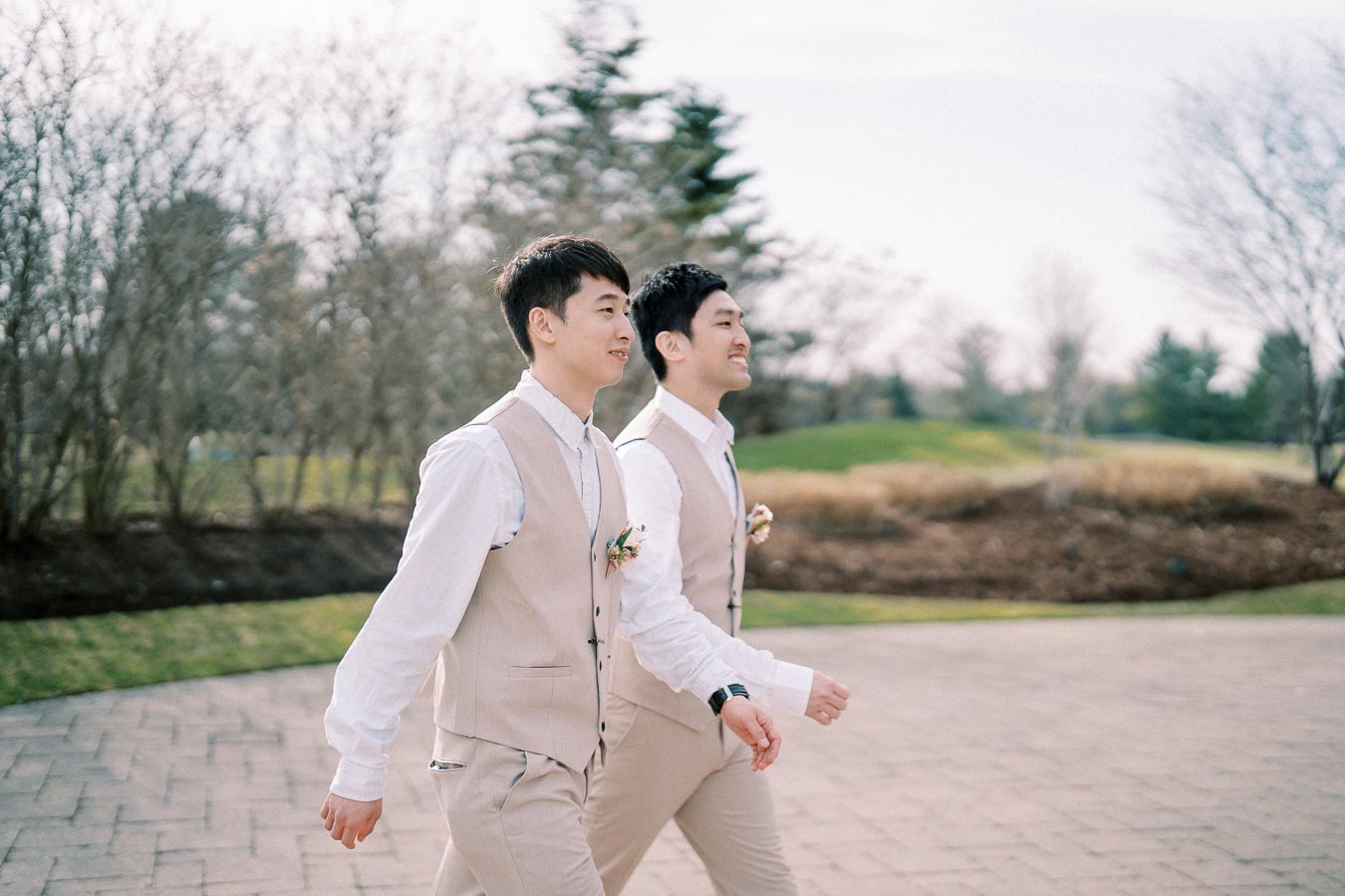 Two groomsmen in beige suits walking outdoors in a garden setting.