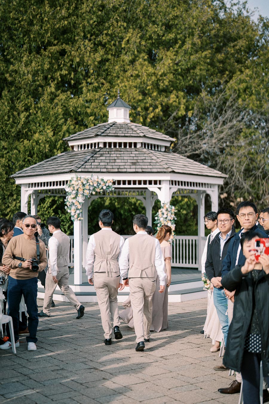 Outdoor wedding ceremony with people gathered near a decorated white gazebo, groomsmen walking towards it in beige suits, surrounded by lush greenery.