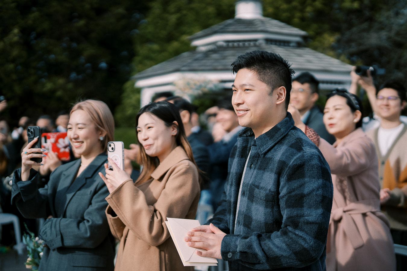 A group of people taking photos and smiling during an outdoor event, with a gazebo in the background, capturing a joyful moment.