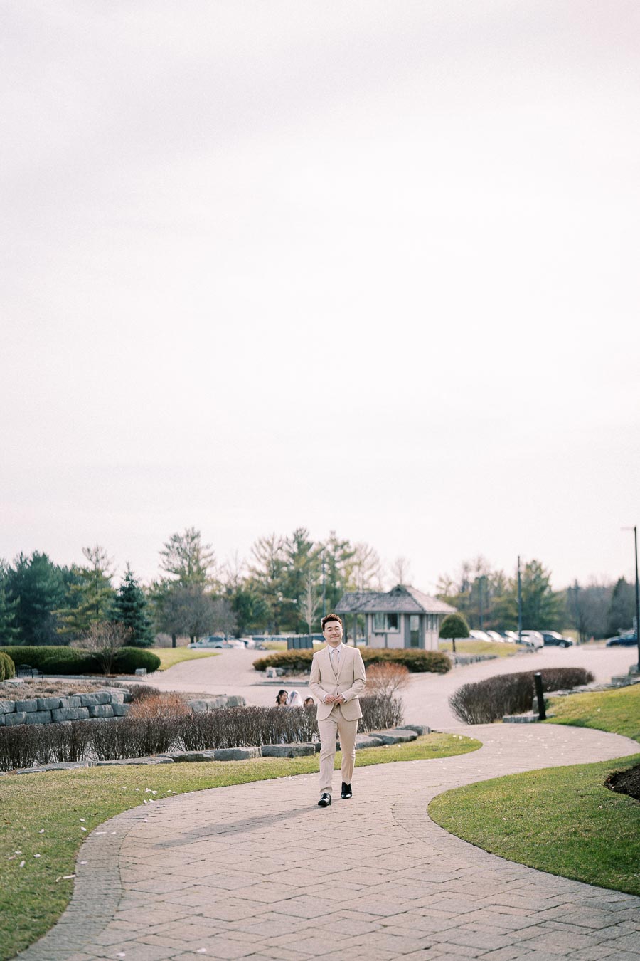 Man in a beige suit walking on a stone path in a park-like setting with trees and a small building in the background.