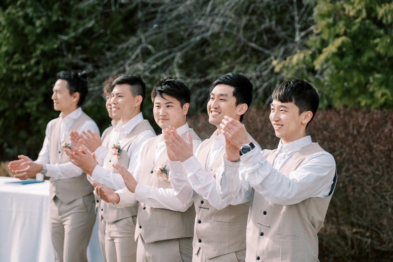A group of groomsmen in beige suits clapping and smiling outdoors at a wedding ceremony, surrounded by greenery.