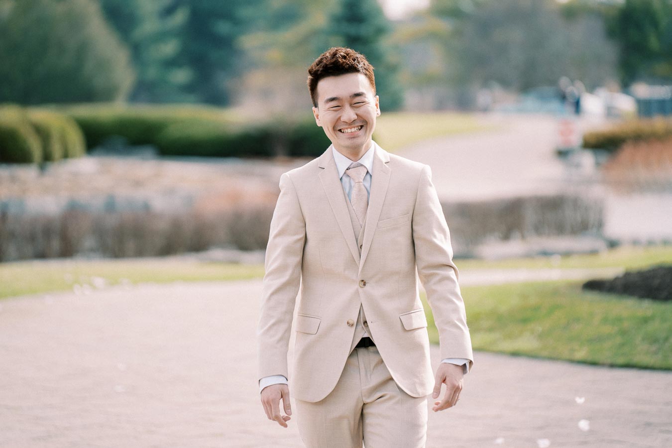 Smiling man in a beige suit walking along a landscaped garden path.