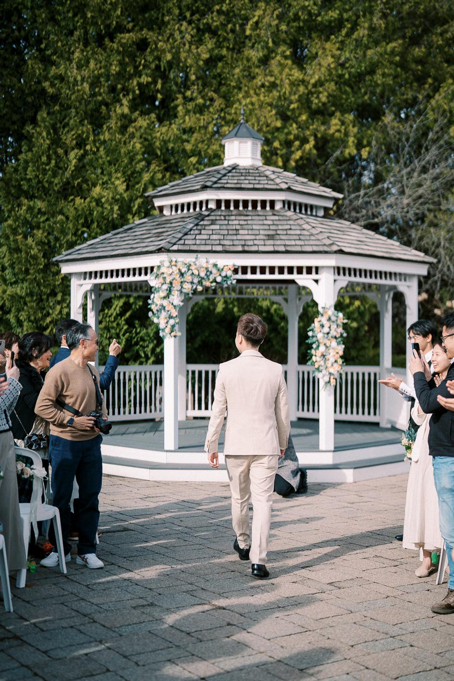 Groom walking towards a decorated gazebo at an outdoor wedding ceremony, surrounded by guests taking photos and cheering.