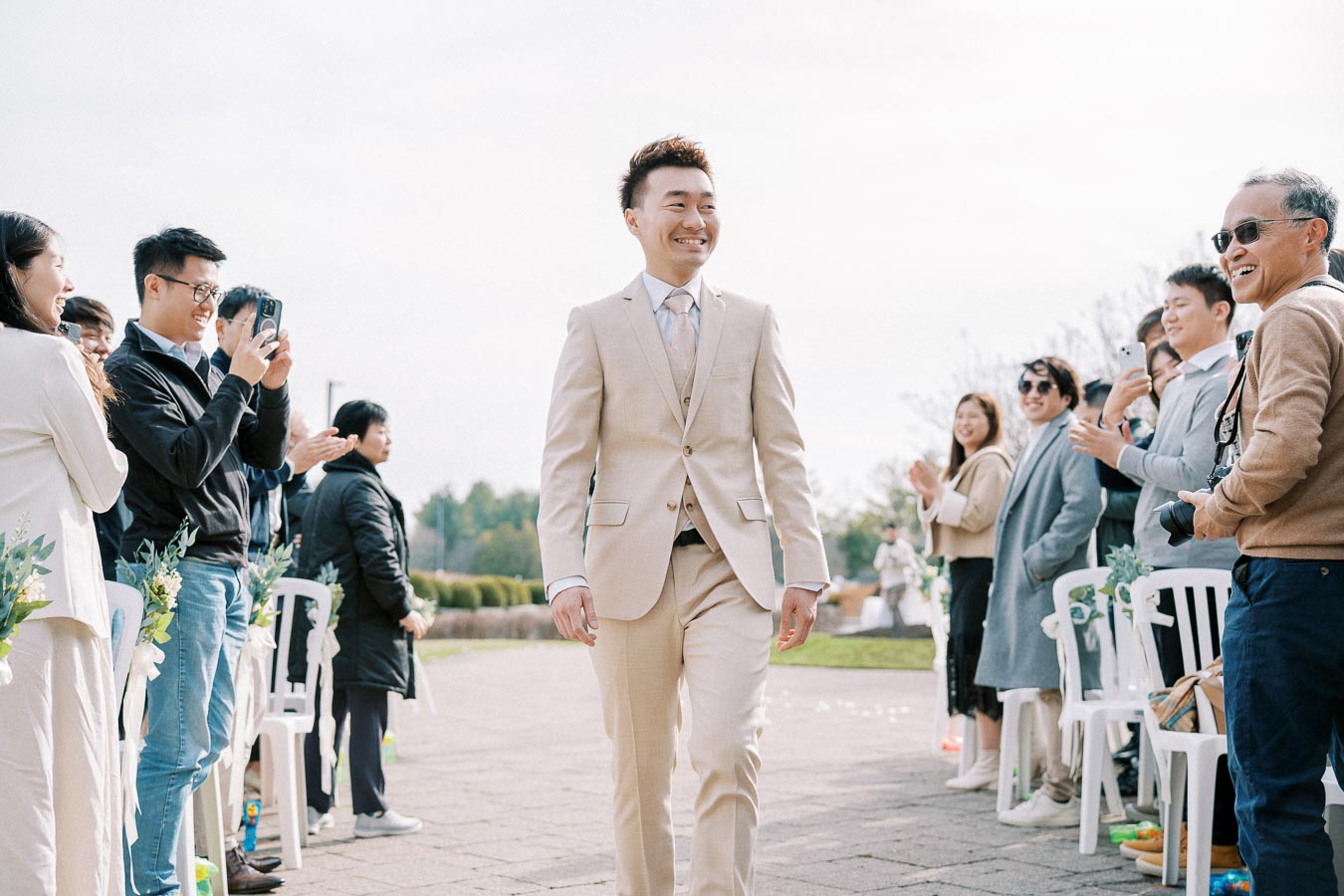 A smiling man in a light beige suit walks down an outdoor aisle, surrounded by applauding and photo-taking guests at a daytime wedding ceremony.