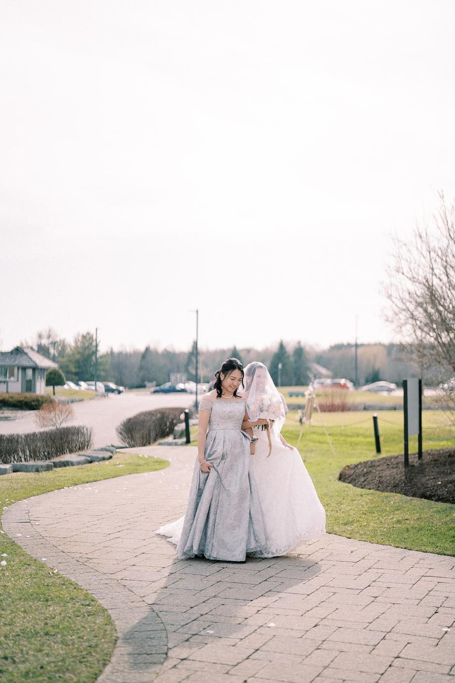 Two women in elegant dresses walking on a stone pathway with greenery in the background, one wearing a bridal gown with a veil, capturing a serene outdoor wedding moment.