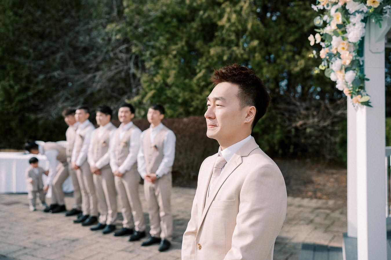 A groom in a light beige suit stands at the altar outdoors, accompanied by groomsmen in matching attire, with lush greenery and floral decorations in the background, capturing a serene wedding moment.
