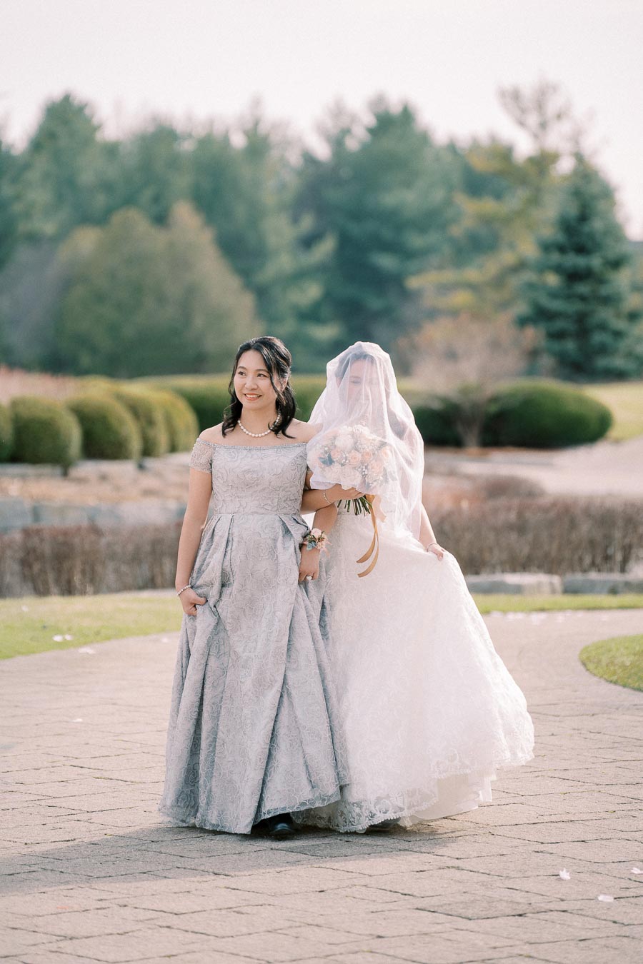 Two women in elegant dresses at an outdoor wedding, with one holding a bouquet and wearing a veil, walking on a stone path surrounded by greenery.