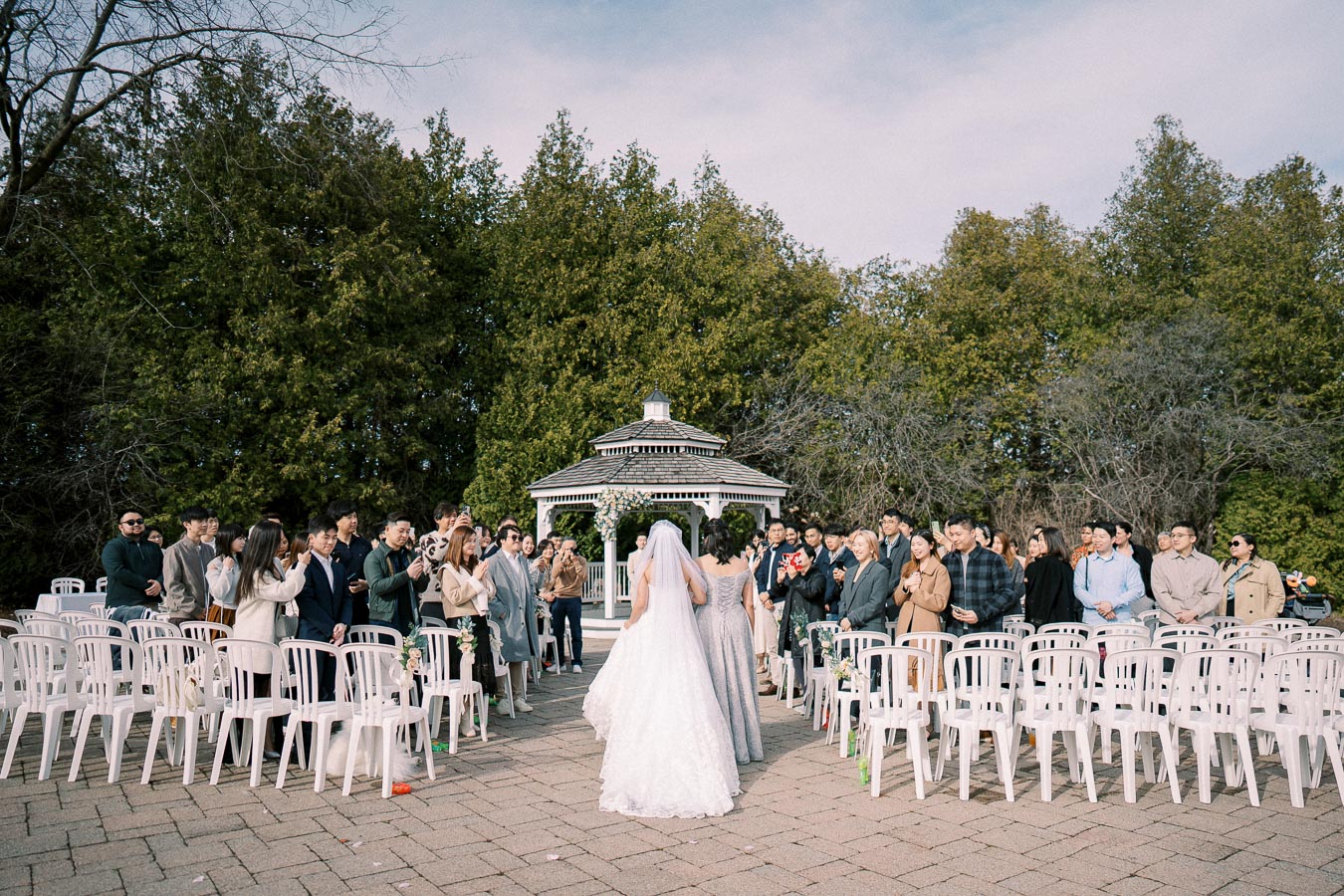 Outdoor wedding ceremony with bride in white gown and veil walking down the aisle toward a decorated gazebo, surrounded by standing guests and lush greenery.