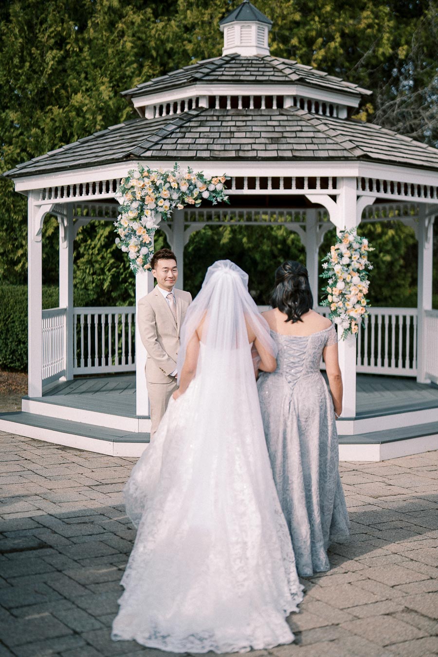 A bride in a white lace gown and veil stands with an older woman in a gray dress, approaching a groom in a beige suit at an outdoor wedding ceremony under a decorated gazebo.