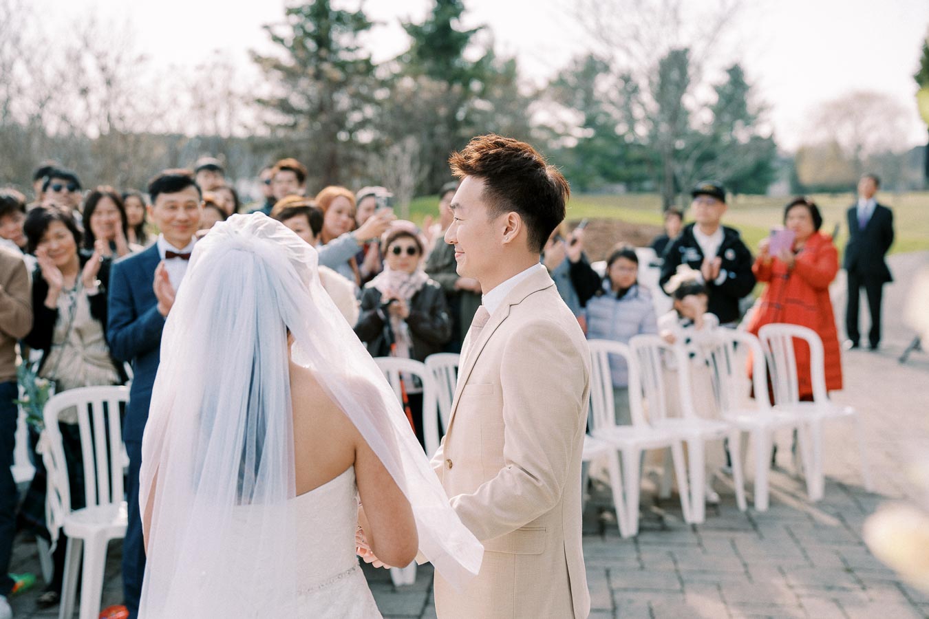 Outdoor wedding ceremony with a bride and groom holding hands, surrounded by smiling guests and trees in the background.