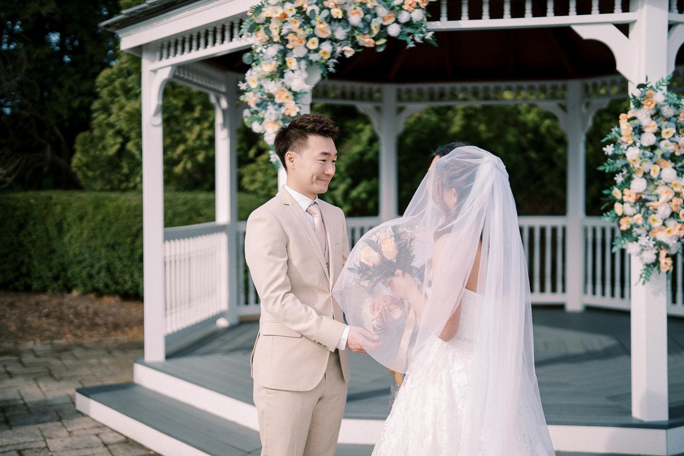 A couple exchanging vows in front of a gazebo adorned with flowers during an outdoor wedding ceremony.