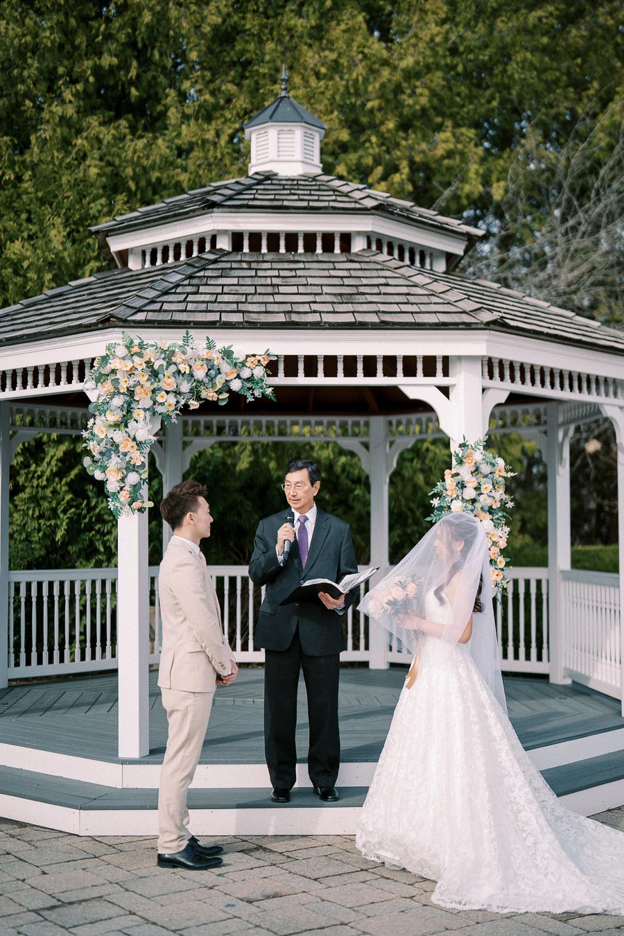 A couple stands under a white gazebo adorned with floral arrangements during an outdoor wedding ceremony, as an officiant in a suit reads from a book.