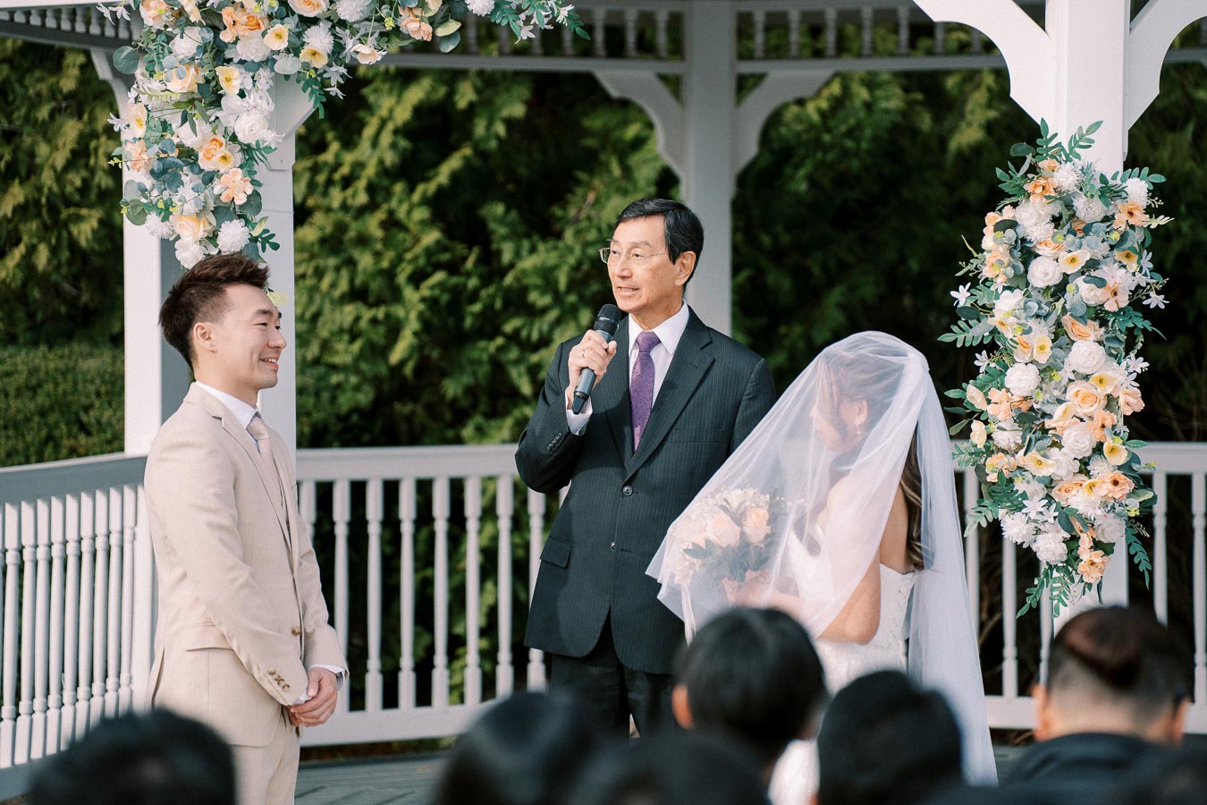Wedding ceremony under a gazebo, featuring a bride in a veil holding a bouquet, a groom in a beige suit smiling, and an officiant speaking into a microphone. Elegant floral arrangements decorate the setting.