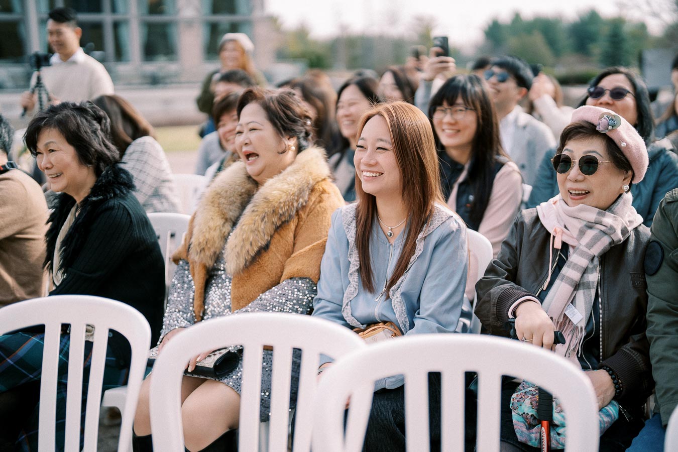 Group of smiling people sitting outdoors on white chairs, enjoying an event on a sunny day, with various expressions of joy and laughter.