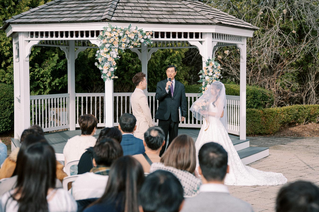 Outdoor wedding ceremony with a couple standing in front of a white gazebo, adorned with floral decorations, as a person officiates. Guests are seated, watching the ceremony on a sunny day.