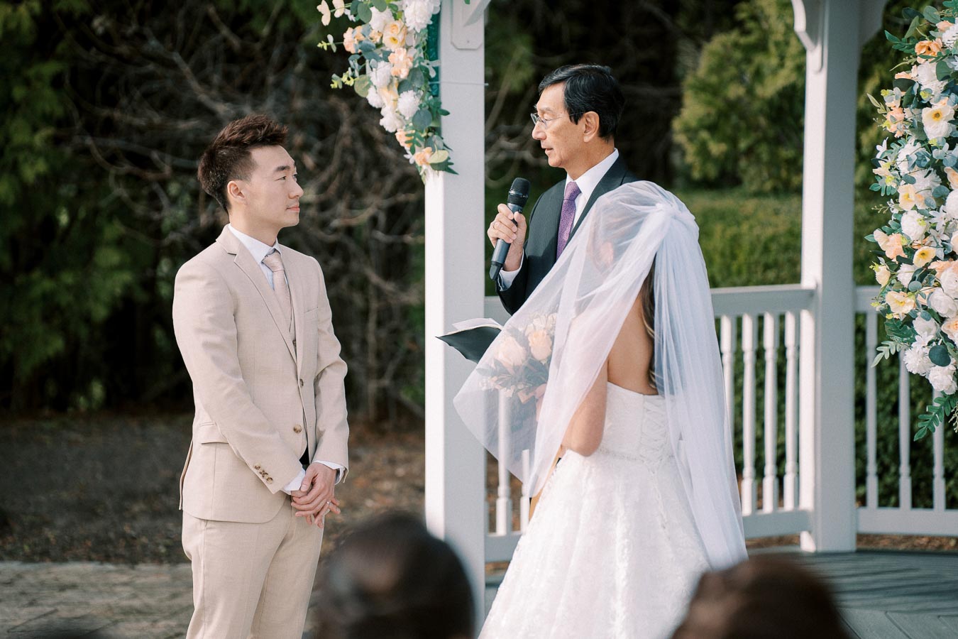Wedding ceremony in an outdoor gazebo with a groom in a beige suit, a bride in a white gown with veil, and an officiant holding a microphone, surrounded by floral decorations.