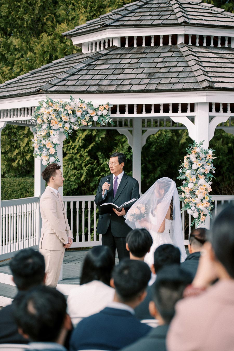 Outdoor wedding ceremony at a gazebo with floral decorations, featuring a couple standing in front of an officiant, surrounded by guests in attendance.