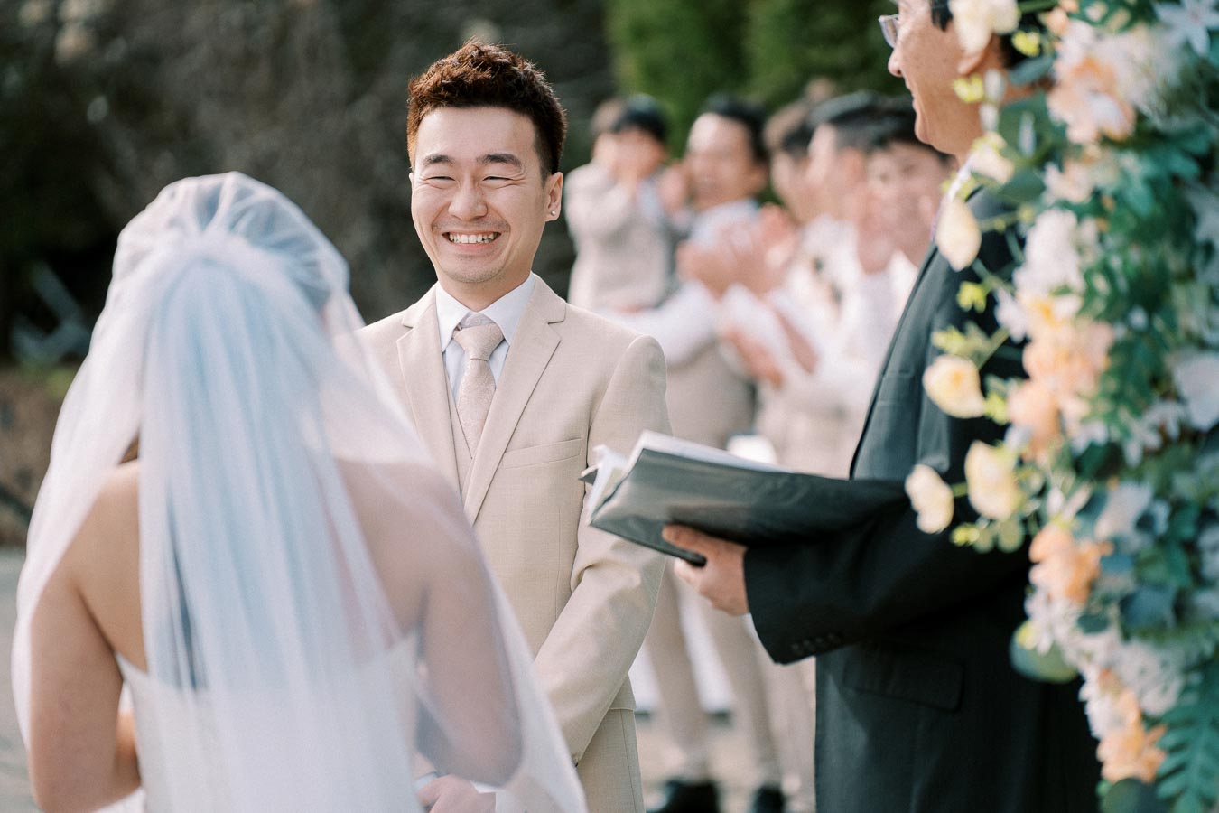 Smiling groom in beige suit standing opposite bride during outdoor wedding ceremony with officiant and floral backdrop.
