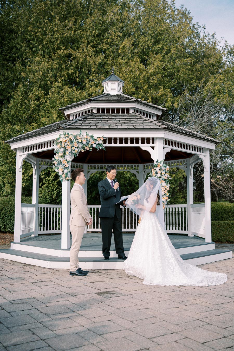 Outdoor wedding ceremony taking place in a beautifully decorated gazebo, with a couple exchanging vows and an officiant presiding, surrounded by lush greenery.