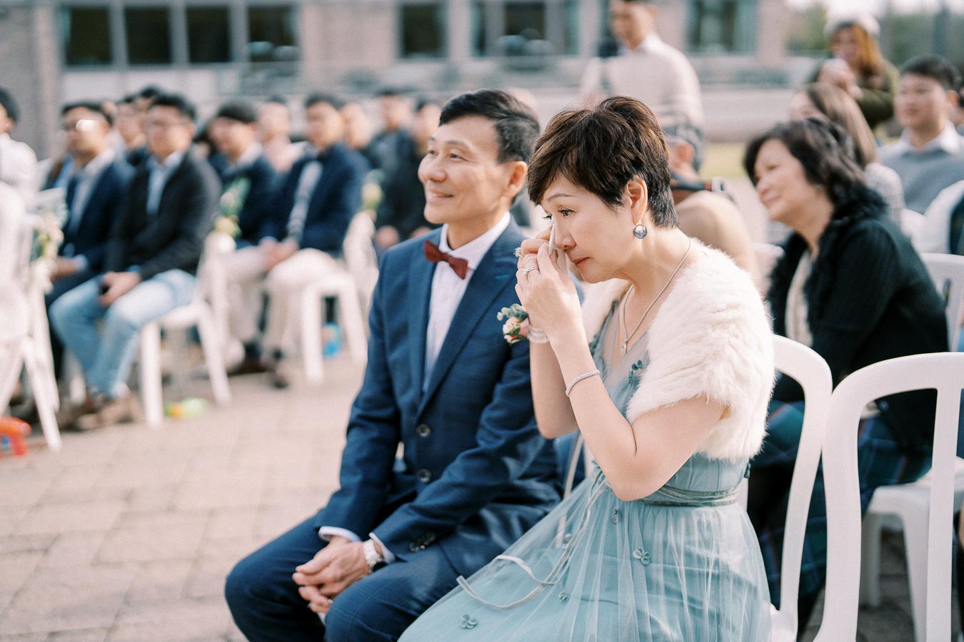 A woman in a teal dress sits beside a man in a navy suit at an outdoor event, visibly emotional. She holds a tissue near her eyes, surrounded by an audience seated on white chairs.