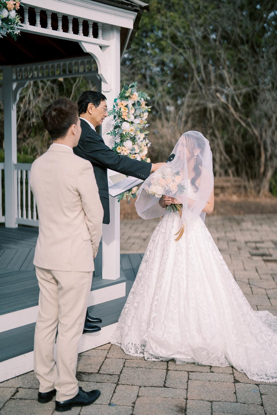 A bride in a white lace wedding gown stands with a bouquet under a gazebo, participating in a wedding ceremony with a groom in a beige suit and an officiant.