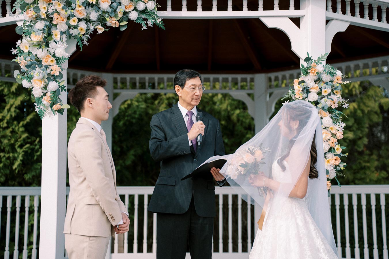 A couple exchanging vows under a floral arch at an outdoor wedding ceremony, with an officiant holding a microphone and guiding the ceremony in a garden gazebo.
