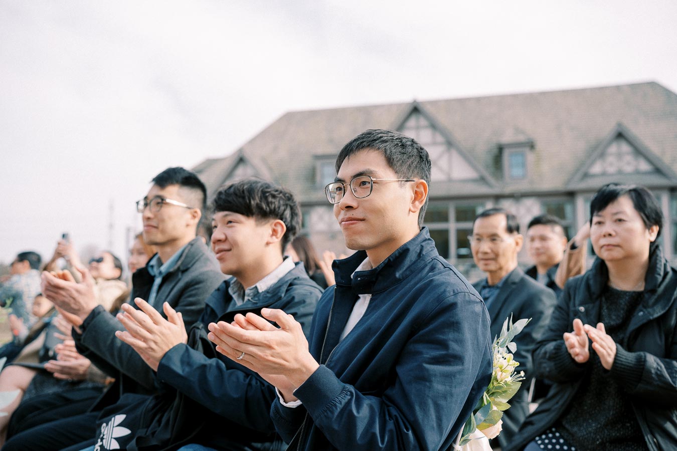 A group of people dressed in formal attire sits outdoors, attentively clapping during an event with a large building in the background.