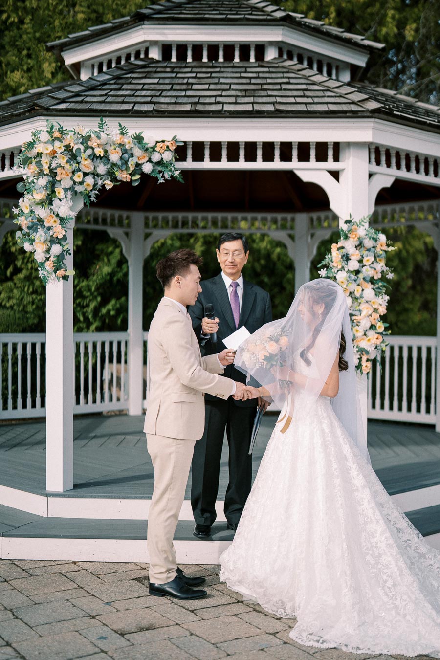 A bride and groom hold hands during their outdoor wedding ceremony at a flower-adorned gazebo, with an officiant standing nearby, in a serene garden setting.