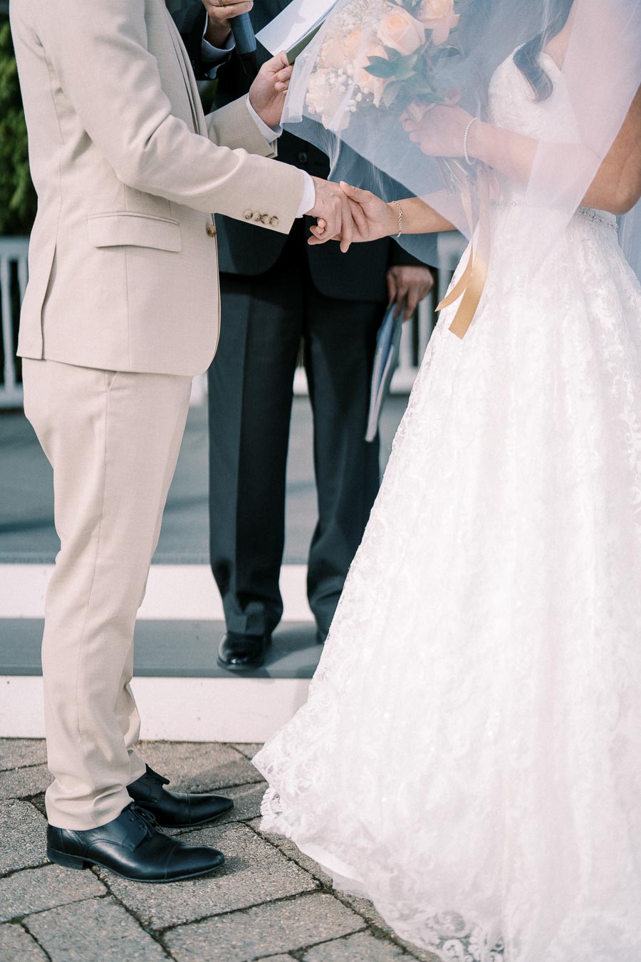 Bride and groom holding hands during outdoor wedding ceremony, dressed in elegant attire, with an officiant in the background.