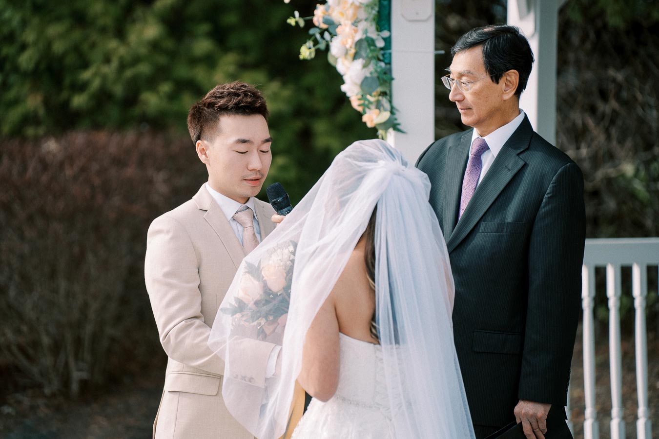 A groom in a beige suit reads vows to his bride in a white wedding dress and veil during an outdoor ceremony, with an officiant in a black suit standing nearby.