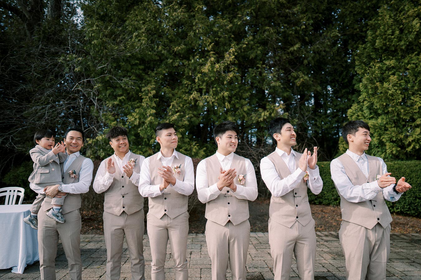 Group of six groomsmen in matching beige suits with boutonnieres, clapping and smiling outdoors next to a table, surrounded by green trees.