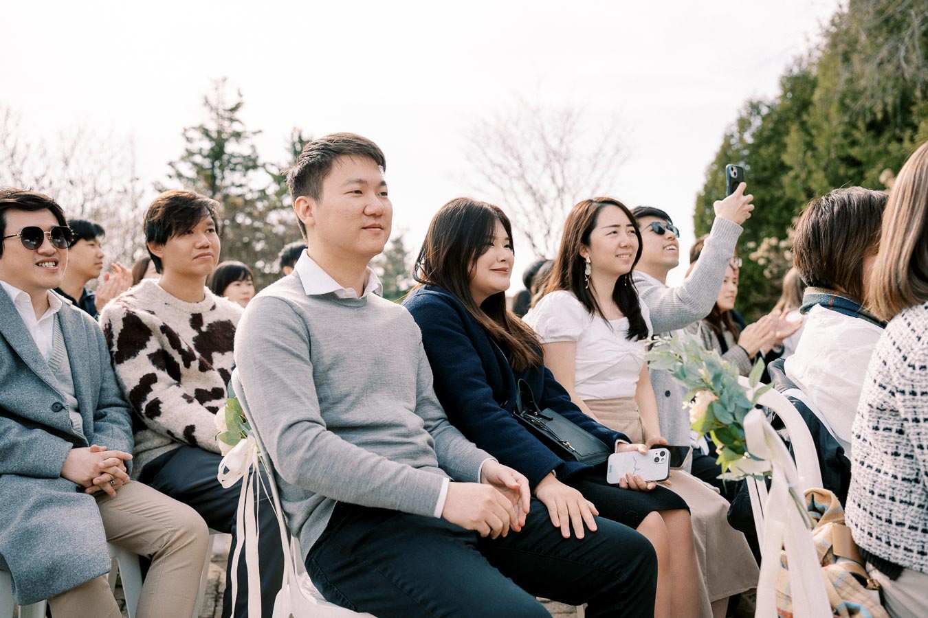 A group of people sitting outdoors at a wedding ceremony, with some guests smiling and using smartphones to capture the moment, surrounded by greenery and trees.