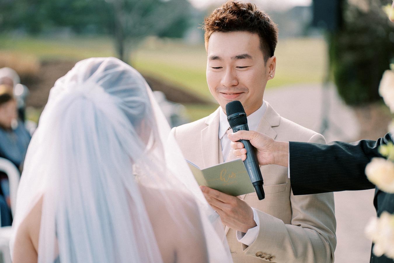 A groom in a beige suit smiles as he reads his vows to the bride during an outdoor wedding ceremony, holding a microphone and a vow booklet marked his.
