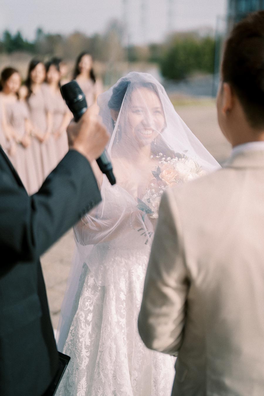 A bride in a lace wedding dress and veil smiles during an outdoor wedding ceremony, holding a bouquet of white and peach flowers. Grooms and bridesmaids in the background create a joyful atmosphere.