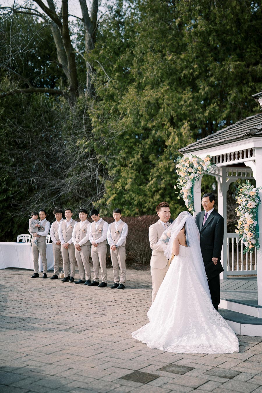 Outdoor wedding ceremony with a groom lifting the bride's veil at a decorated gazebo, surrounded by groomsmen in matching suits, under lush green trees.