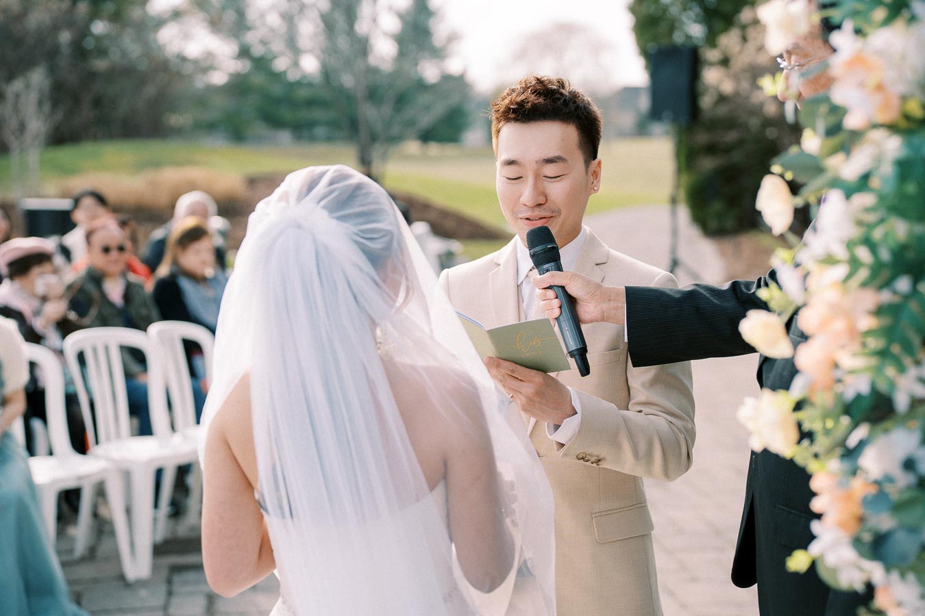 Bride and groom exchanging vows during an outdoor wedding ceremony, with the groom holding a microphone and reading from a card, surrounded by guests and floral decorations.
