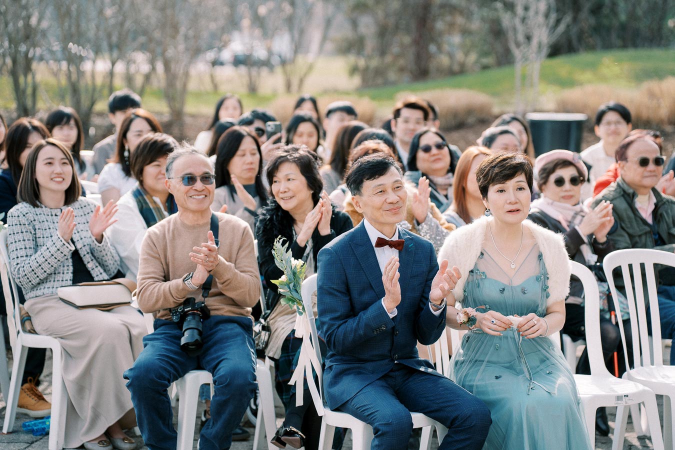 A diverse group of people seated outdoors, dressed in formal attire, clapping and smiling during a daytime event, surrounded by greenery and natural light.