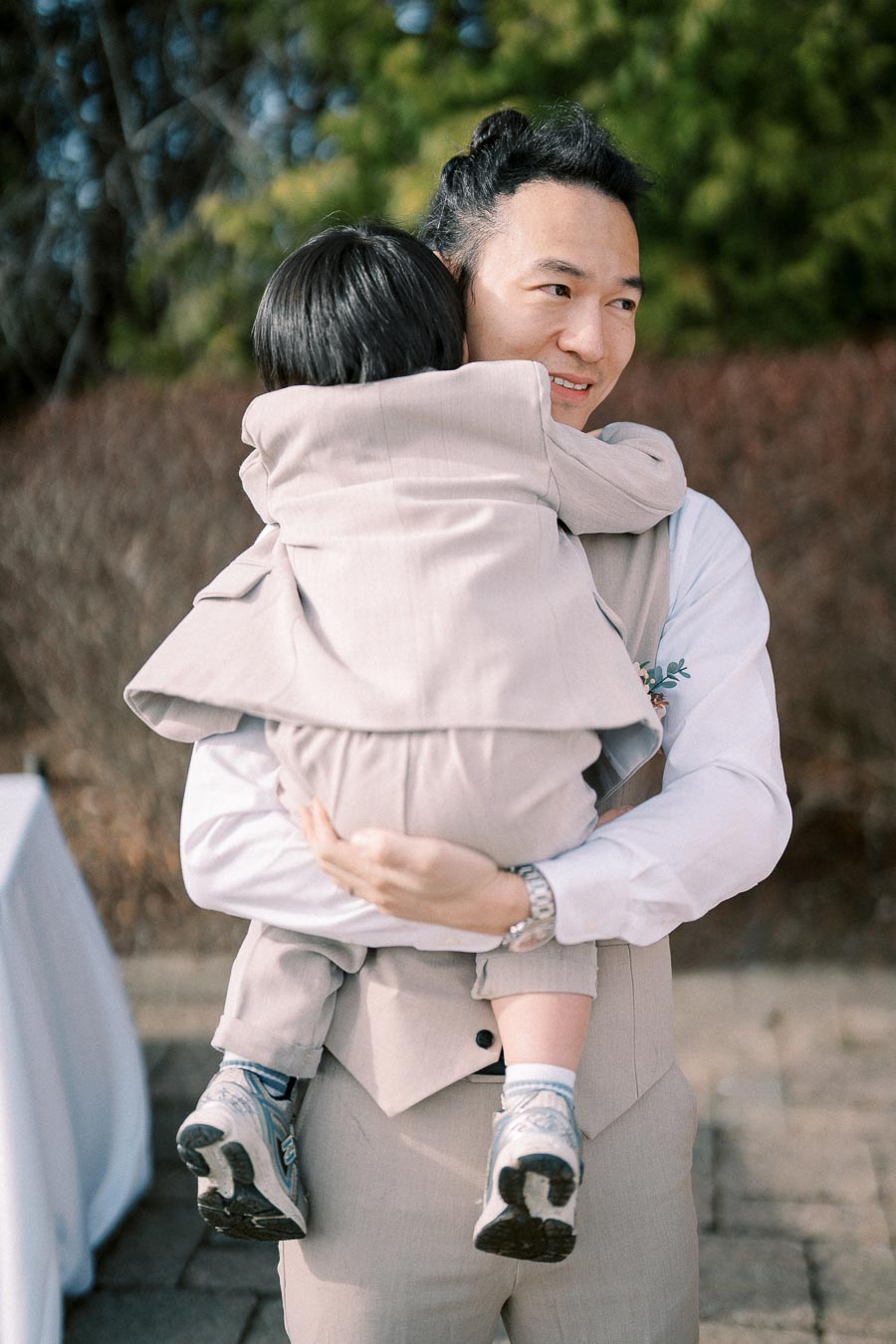 Father holding child outdoors at a wedding, both in light-colored suits, with trees in the background.