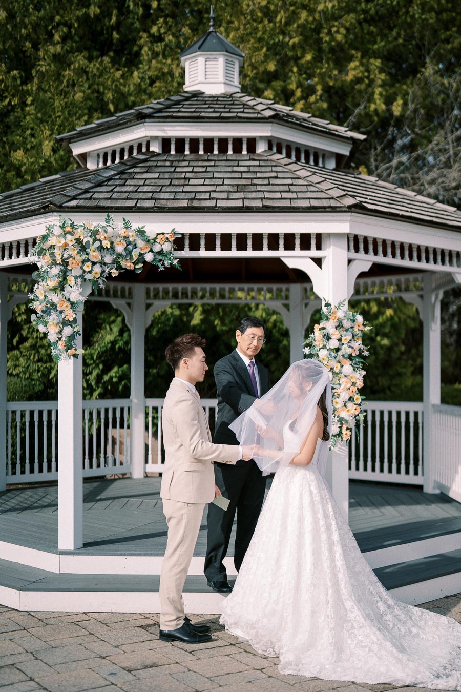 Outdoor wedding ceremony at a beautifully decorated gazebo with a bride in a white gown and veil holding hands with the groom in a beige suit, as officiant looks on.